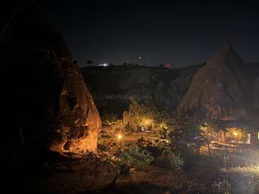 This nighttime image shows a cozy outdoor area illuminated by warm lights, surrounded by unique rock formations and trees, creating a serene, inviting atmosphere under a dark sky.