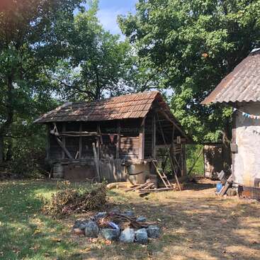 Une vieille cabane en bois avec un toit en tuiles se trouve au milieu d'arbres verts. Devant, il y a un foyer en pierre et des branches éparpillées sur l'herbe ensoleillée.
