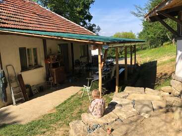 A small child sits on stone steps outside a rustic house with a tiled roof, surrounded by greenery, garden tools, and a covered outdoor dining area.