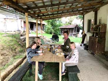 Four people enjoy a meal together at a rustic outdoor table under a wooden roof, surrounded by greenery, wooden structures, and a cozy, countryside setting.
