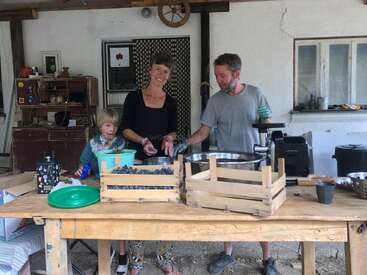 A family stands around a wooden table processing fresh blueberries. Wooden crates, bowls, and kitchen equipment are visible, creating a cozy, rustic atmosphere in an outdoor setting.