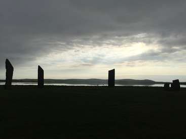 The image depicts a serene landscape featuring a body of water, a grassy field, and a row of stone monoliths set against a cloudy sky with distant hills.