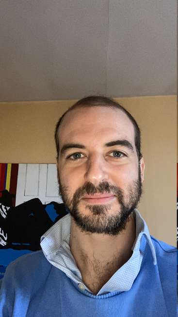 The image depicts a man with short dark hair and a beard, wearing a blue sweater over a collared shirt, standing in front of a wall with a colorful painting.