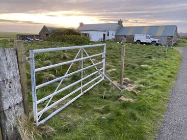 The image depicts a serene rural landscape featuring a white gate, a stone house, and a truck, set against a backdrop of a cloudy sky at sunset.