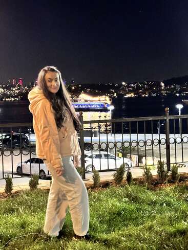 A woman stands on green grass by a fence at night, with a brightly lit boat and city skyline illuminating the water in the background.