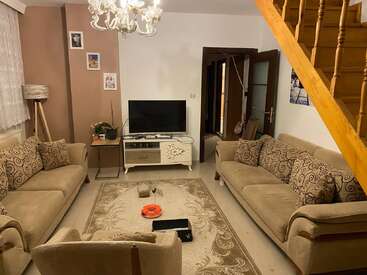 This cozy living room has beige sofas, a patterned rug, a TV, wooden stairs, decorative pillows, framed photos, plants, a chandelier, and warm lighting.