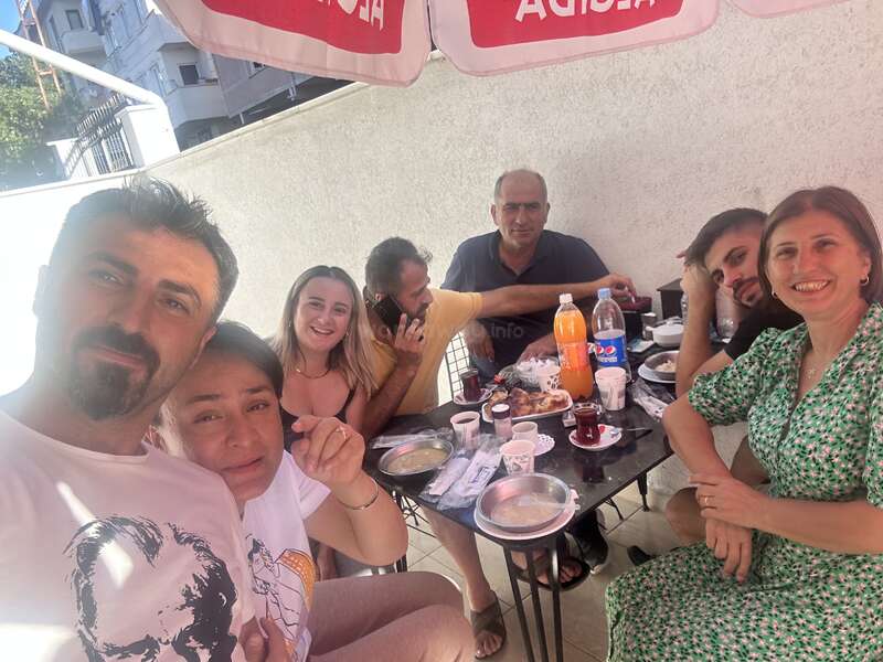 A group of seven people enjoys an outdoor meal together. They are smiling, drinking tea, and seem happy, gathered around a table with food and drinks.