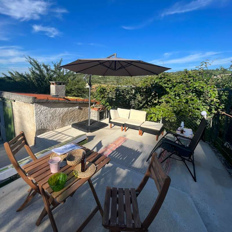 This cozy outdoor patio features wooden furniture, a sofa, a large umbrella, drinks, and lush greenery under a bright blue sky—perfect for relaxing and socializing.