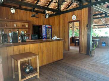 This image shows a rustic wooden bar with shelves of bottles, coffee machines, a beverage fridge, wooden furniture, a clock, ceiling fans, and natural light.