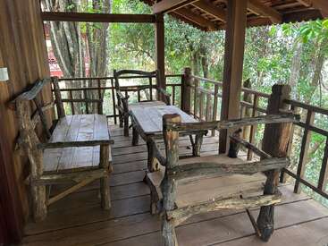 A rustic wooden patio features handmade benches and a table, surrounded by wooden railings and lush green trees, creating a peaceful, natural outdoor seating area.