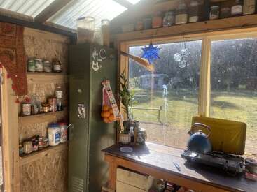 Sunlit rustic kitchen with wooden shelves, jars, metal locker, hanging oranges, blue kettle on stove, potted plant, and large window overlooking a green outdoor yard.