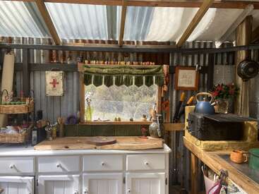 This cozy rustic kitchen features corrugated metal walls, white cabinets, wooden counters, a blue kettle, baskets, utensils, plants, green curtains, and sunlight streaming through the roof.