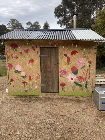A small shed with a corrugated metal roof features a wooden door and vibrant flower mural. Surrounded by trees, string lights hang above the entrance.