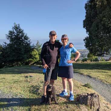 A smiling couple stands outdoors on a grassy hill, embracing each other, with a small black dog sitting in front. Trees and a scenic view behind them.