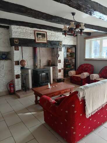 Essa sala de estar aconchegante tem vigas expostas, lareira de tijolos caiados, lareira a lenha, sofás com estampa vermelha, mesa de centro de madeira, estante de livros, decoração vintage e luz natural.