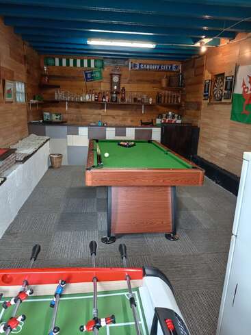 Esta acogedora sala de juegos cuenta con una mesa de billar, un futbolín, un tablero de dardos, una zona de bar con botellas, recuerdos deportivos, paredes de madera, techo azul y una bandera galesa.