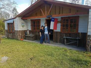 A rustic wooden house with two people standing at the entrance, a Chilean flag hanging, and green grass in front. The atmosphere feels peaceful and welcoming.