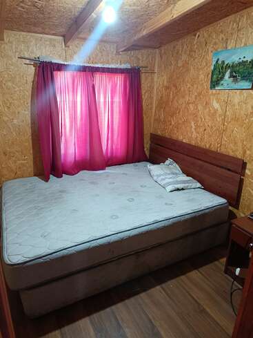 A cozy wooden room with a mattress bed, single pillow, maroon curtains covering the window, a glowing ceiling light, a small nightstand, and a painting.