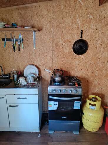 A simple kitchen with wooden walls, a stove, gas cylinder, dish rack, knives on a magnetic strip, a shelf, a frying pan, and a kettle.