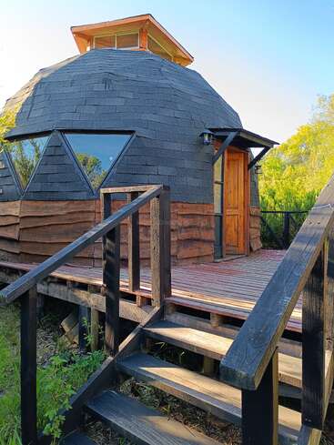 A unique dome-shaped cabin with a shingled roof, wooden door, angled windows, and elevated wooden porch, surrounded by greenery under a clear blue sky.