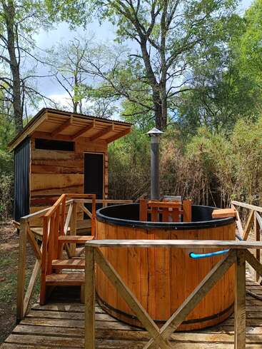 A rustic wooden hot tub and sauna sit outdoors on a wooden deck, surrounded by lush trees and greenery, creating a relaxing, natural retreat in nature.
