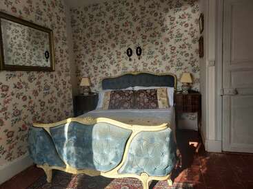 Esta imagen muestra un dormitorio vintage con papel pintado floral, una cama tapizada en azul, cabecero y piecero ornamentados, mesitas de noche de madera, lámparas clásicas y cuadros decorativos.