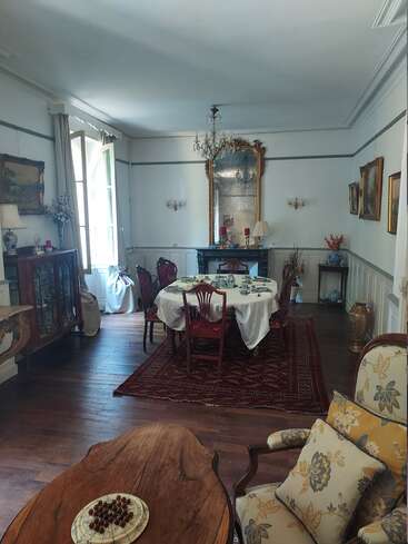 Comedor elegantemente decorado con muebles vintage, lámpara de araña, espejo grande y cuadros. Mesa preparada para el té, suelos de madera, sillón floral, ambiente acogedor y atractivo.