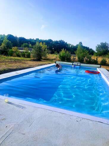 Una persona se relaja en una piscina exterior resplandeciente, rodeada de vegetación y árboles. Hay una bebida junto a la piscina, con un cielo azul sobre ella. Ambiente tranquilo.