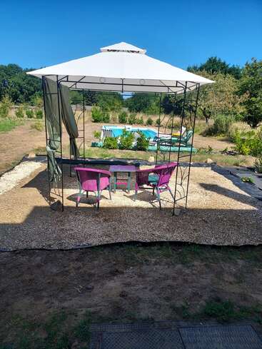 Una escena de jardín muestra una glorieta blanca con sillas moradas y una mesa debajo, con vistas a una pequeña piscina, rodeada de vegetación y un cielo azul claro.