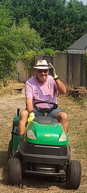 Un hombre con sombrero, gafas de sol, camisa rosa, guantes y pantalones cortos conduce un tractor cortacésped verde en un día soleado, rodeado de árboles y arbustos.
