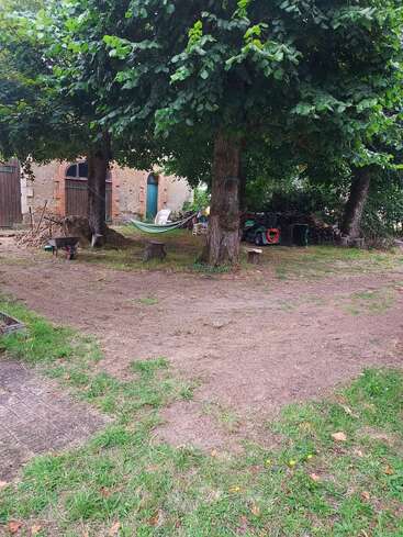 Esta imagen muestra un tranquilo patio trasero con árboles, una hamaca, un banco, una carretilla, leña apilada, herramientas de jardín esparcidas y una casa rústica al fondo.