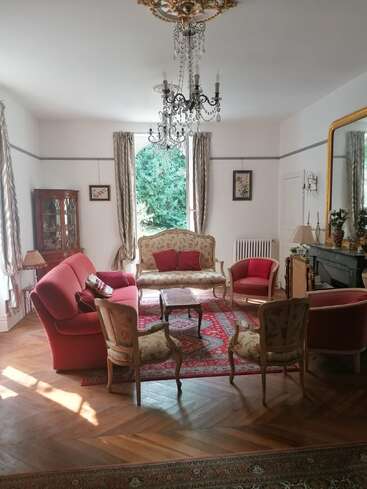 Esta elegante sala de estar cuenta con muebles vintage, detalles en rojo, una lámpara de araña ornamentada, un gran espejo, suelos de madera, obras de arte clásicas, ventanas altas y un ambiente cálido y acogedor.
