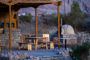 Uma área de estar ao ar livre com mesas de madeira, cadeiras de vime e forno de pedra, em um ambiente rústico e natural cercado por árvores e montanhas, oferece tranquilidade.