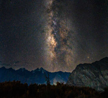Um céu noturno hipnotizante com a Via Láctea se eleva acima de picos de montanhas escarpadas e silhuetas escuras, criando uma paisagem natural de tirar o fôlego sob um manto de inúmeras estrelas.