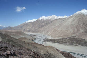A imagem retrata uma paisagem vasta e árida com o leito de um rio, montanhas cobertas de neve e um céu azul claro, evocando uma sensação de beleza natural e tranquilidade.