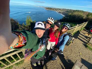 L'image montre quatre personnes posant pour un selfie sur une falaise surplombant l'océan, avec une clôture en bois et de la verdure en arrière-plan.