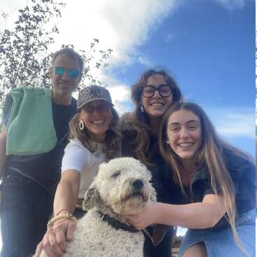 Eine glückliche vierköpfige Familie posiert draußen unter blauem Himmel, strahlt über das ganze Gesicht und hält liebevoll ihren flauschigen weißen Hund im Arm. Bäume und Wolken sorgen für eine natürliche Kulisse.