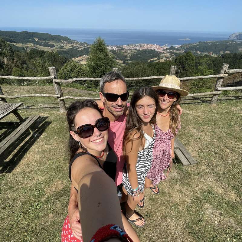L'image représente une famille de quatre personnes posant pour un selfie dans un cadre extérieur pittoresque, avec une vue panoramique de collines ondulantes et d'un plan d'eau en arrière-plan.