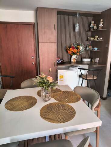 Un acogedor comedor con una mesa blanca, cuatro sillas, manteles individuales dorados y un jarrón con flores. Unas estanterías decorativas y una pequeña barra completan el conjunto.