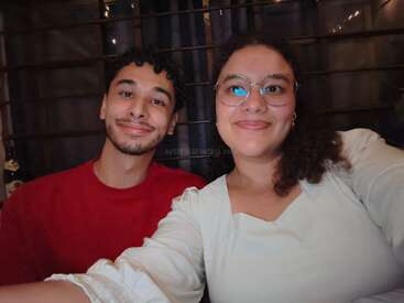 Un hombre y una mujer sonrientes se hacen un selfi en un interior. El hombre lleva una camisa roja; la mujer, gafas y ropa blanca. Al fondo se ve una rejilla metálica y unas luces.