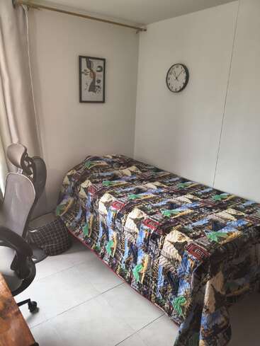 Una habitación pequeña y ordenada con una cama individual cubierta por una colcha de colores, una silla de oficina, cuadros en la pared, un reloj, cortinas y suelo de baldosas.