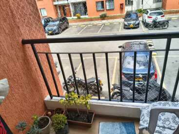 Un pequeño balcón con plantas en macetas da a un aparcamiento lleno de coches y motos. La zona está rodeada por una red de seguridad y una barandilla metálica.