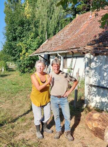 The image depicts a man and woman standing outside a rustic white building with a brown tiled roof, surrounded by trees and grass.