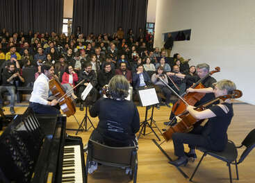 A quartet of musicians performs for an attentive audience in a modern hall. The audience sits closely together, engaged and enjoying the live classical music performance.