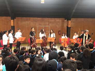 A group of musicians with string instruments performs in front of a large audience of children in a hall with wooden walls and red chairs.
