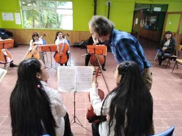 In a brightly colored classroom, students play string instruments while a teacher leans in to guide two girls with music sheets, fostering a musical learning environment.