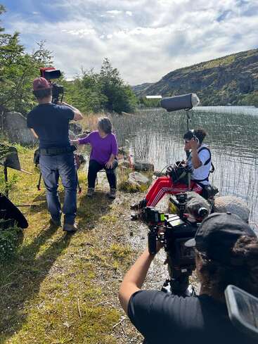 A film crew records an interview outdoors by a lake, surrounded by mountains. Cameras, microphones, and equipment are visible, with bright skies and lush scenery.