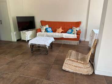 La photo montre un salon chaleureux avec un canapé orange, des coussins décoratifs, une télévision, un fauteuil en osier, des petites tables recouvertes d'une nappe et un sol carrelé.