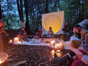 A group of people gathers in a forest clearing at dusk, surrounded by lanterns, playing music and singing. A decorative tapestry hangs behind the performers.