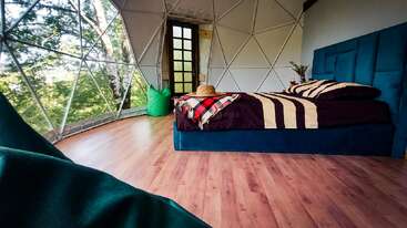 Cette image montre une chambre dôme confortable et moderne avec de grandes fenêtres, un lit bleu, un plancher en bois, un pouf vert, une vue sur la nature et la lumière naturelle qui pénètre à l'intérieur.